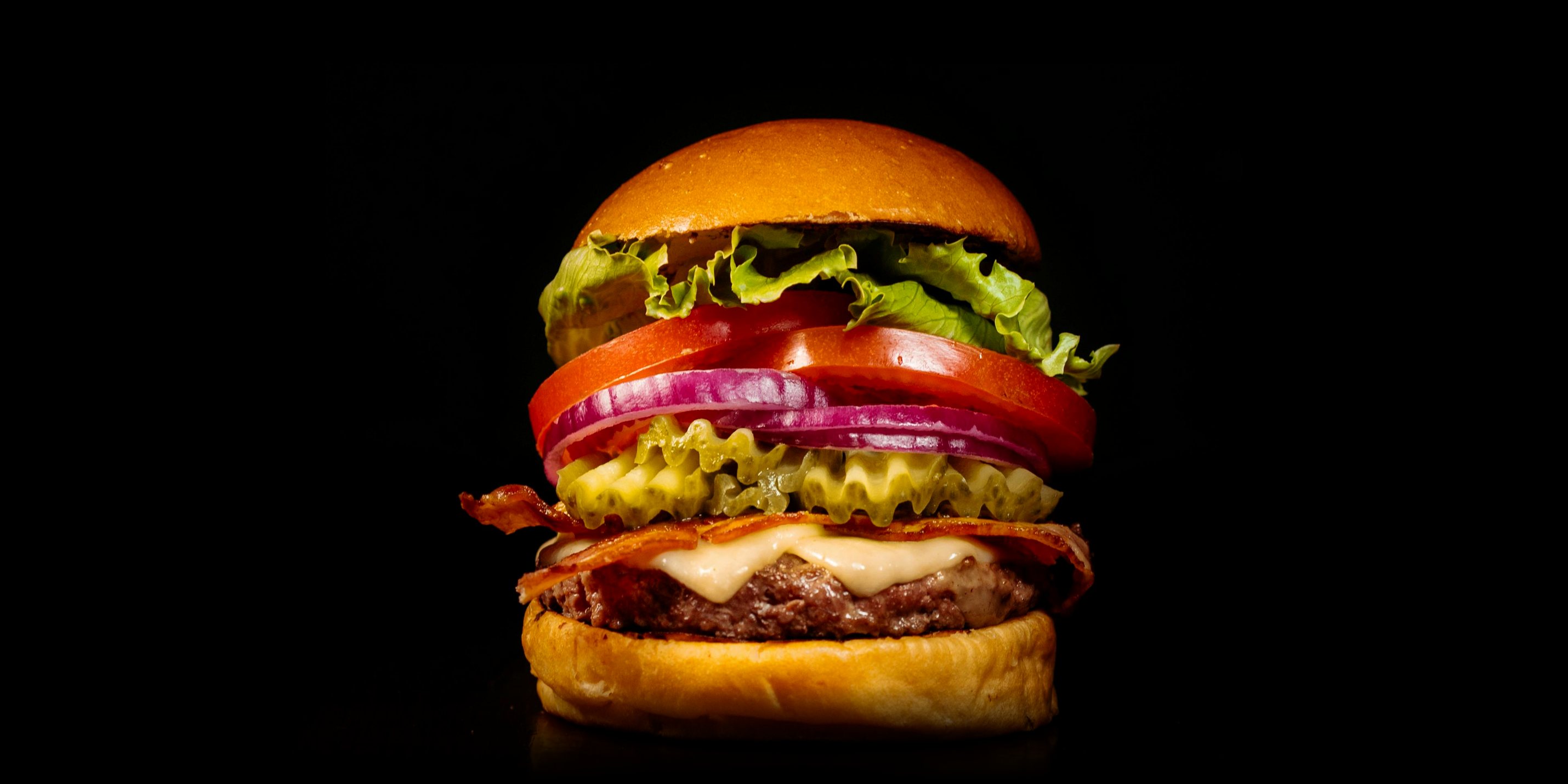 A burger built in layers: bun, lettuce, tomato, red onion, pickles, bacon, cheese and patty.